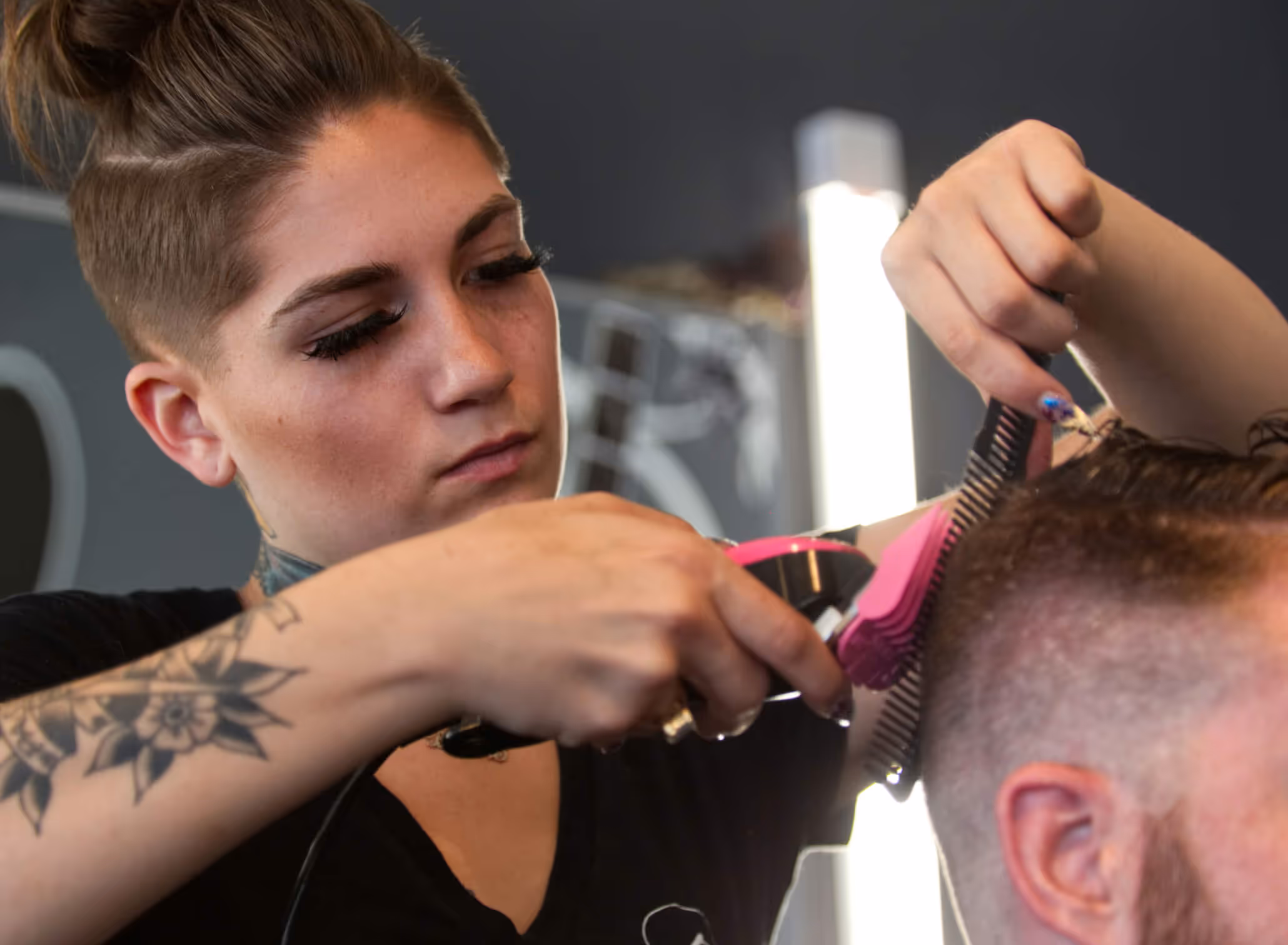 Tattooed hairstylist with short hair and eyelashes cutting a man's hair using clippers and a comb in a barber shop.