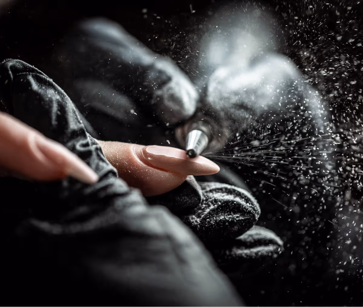 Close-up of a gloved hand using an electric nail drill on a manicured fingernail with dust particles in the air.