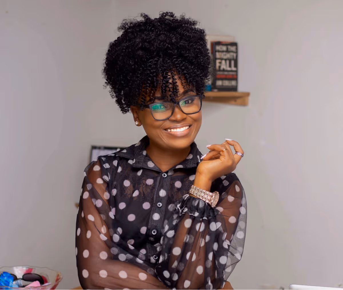 Smiling woman with curly hair, glasses, and a black polka-dot sheer blouse posing indoors.