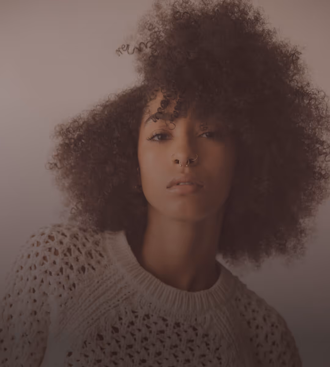 Close-up of a person with voluminous curly hair, nose rings, and a cream-colored knitted sweater.