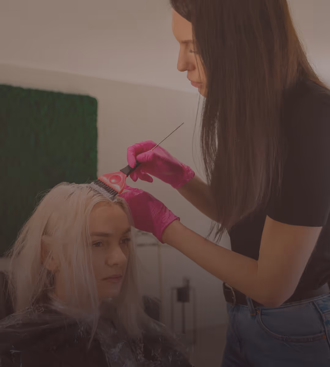 Hairdresser wearing pink gloves applying hair dye to a woman's blonde hair using a brush in a salon.