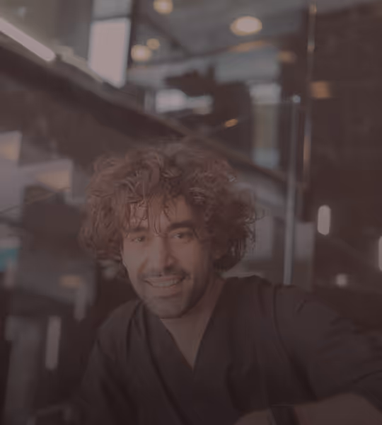 Smiling man with curly hair wearing a black shirt inside a modern building with glass and lights.