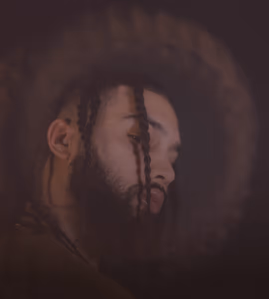 Close-up side profile of a man with braided hair, a beard, and a dark, blurred circular background.