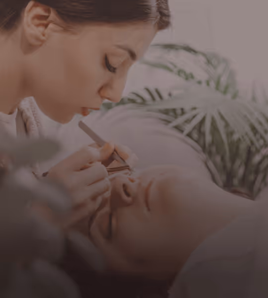 Woman applying eyelash extensions to a client lying down with closed eyes in a serene setting.