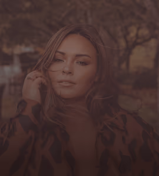 Portrait of a woman with long hair, wearing a patterned jacket, outdoors in a softly blurred natural setting.