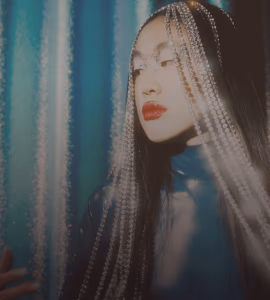 Woman with long dark hair wearing sparkling beaded strands over her head and bright red lipstick, against a blurred blue background.