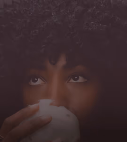 Person with curly hair holding and drinking from a white cup, looking upward.