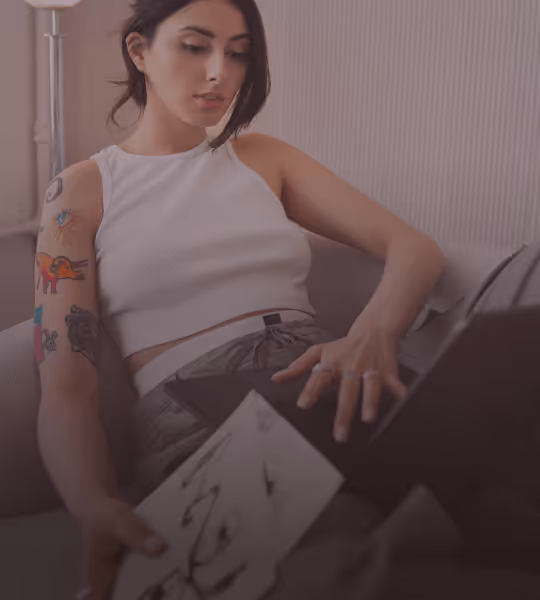 Young woman with tattoos on her arm sitting on a couch, holding tattoo design sheets and working on a laptop.