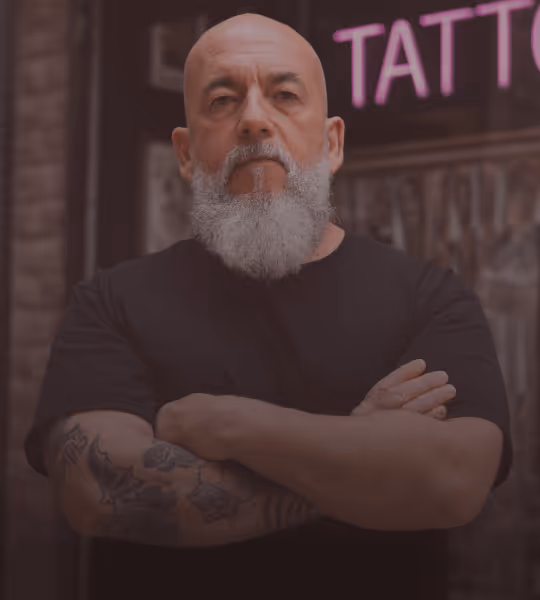 Bald man with a gray beard and tattooed arms crossing his arms in front of a tattoo shop sign.