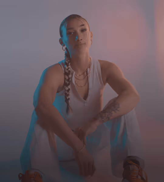 Young woman with braided hair sitting cross-legged wearing a sleeveless top and layered necklaces in soft lighting.