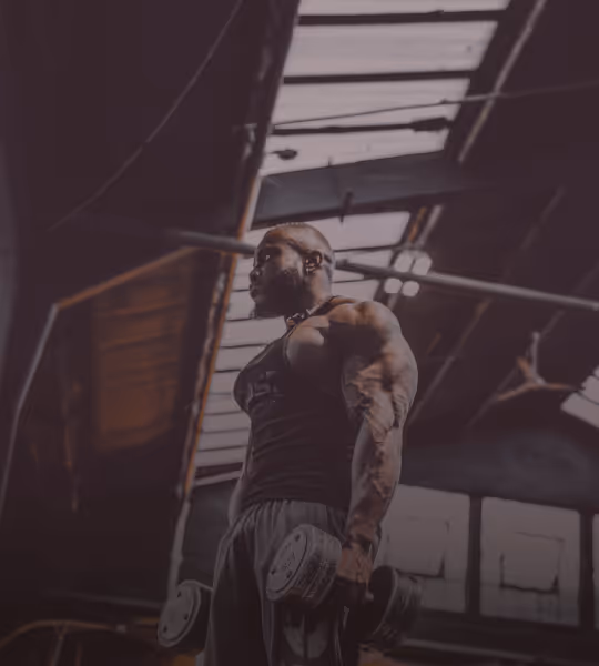 Muscular man wearing headphones lifting dumbbells in a gym with large skylights.