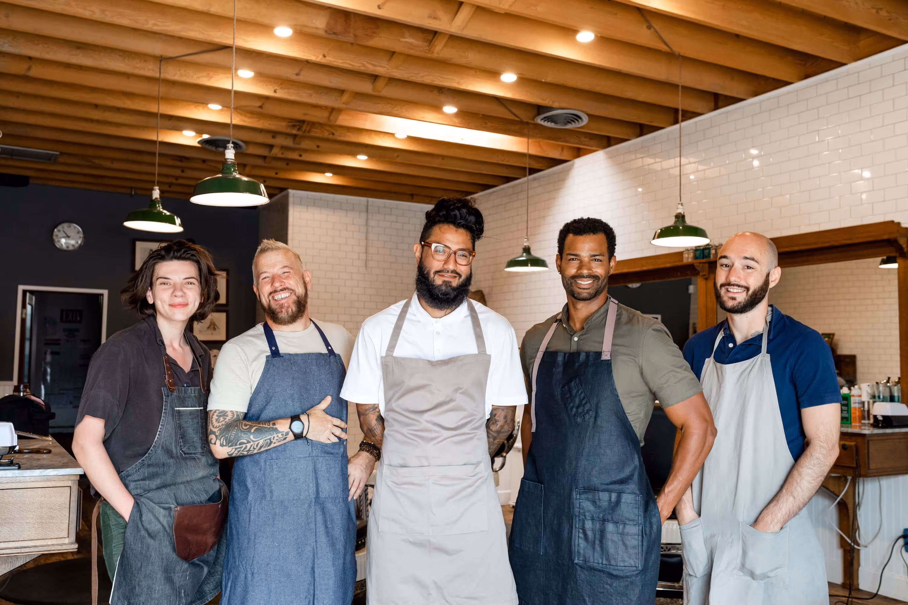 Five diverse barbers wearing aprons smiling and standing in a modern barbershop with wooden ceiling and white tiled walls.