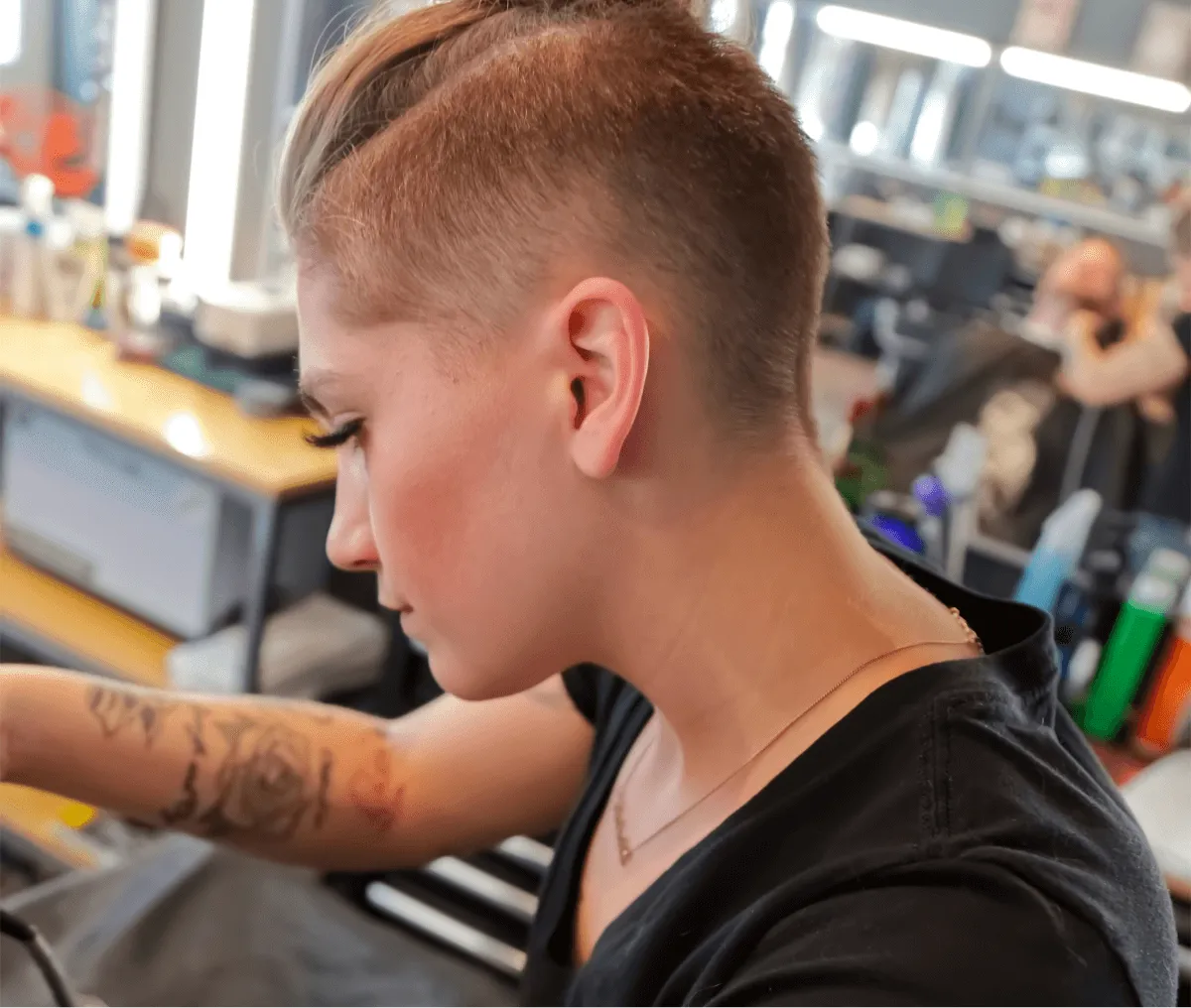 Person with short hair and tattooed arm focused on a task in a busy barbershop.
