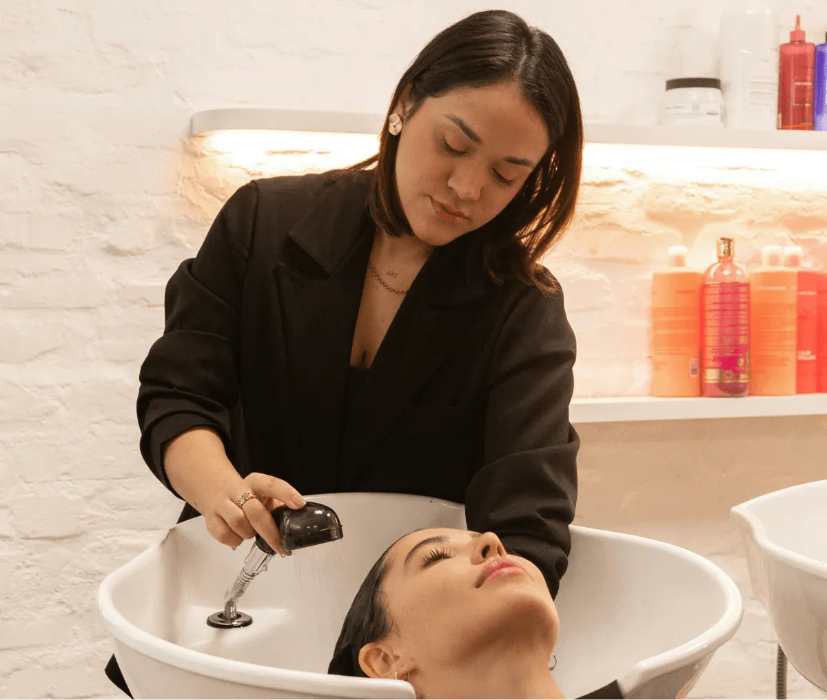 Woman washing another woman's hair over a salon sink with haircare products on shelves behind.