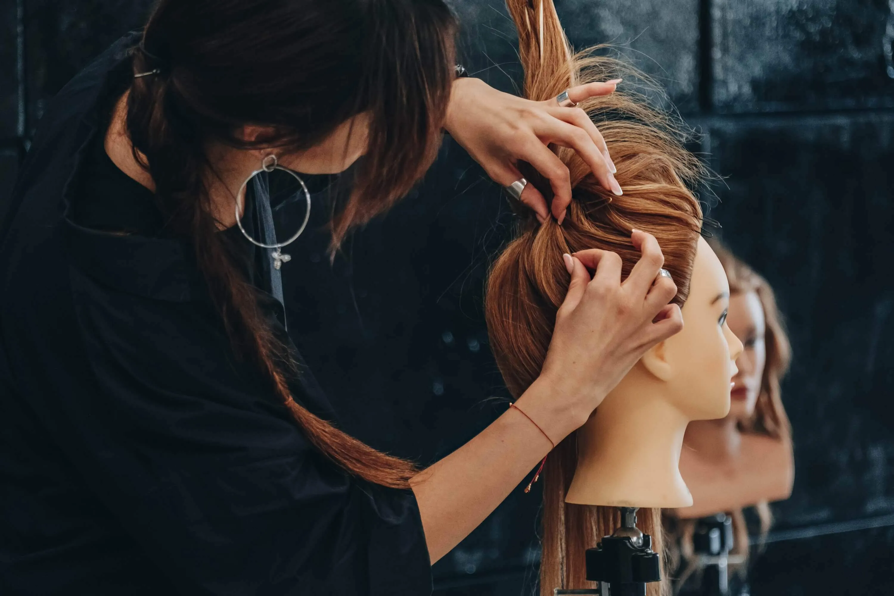 Hairstylist styling a red-haired mannequin head in a salon with another mannequin head blurred in the background.
