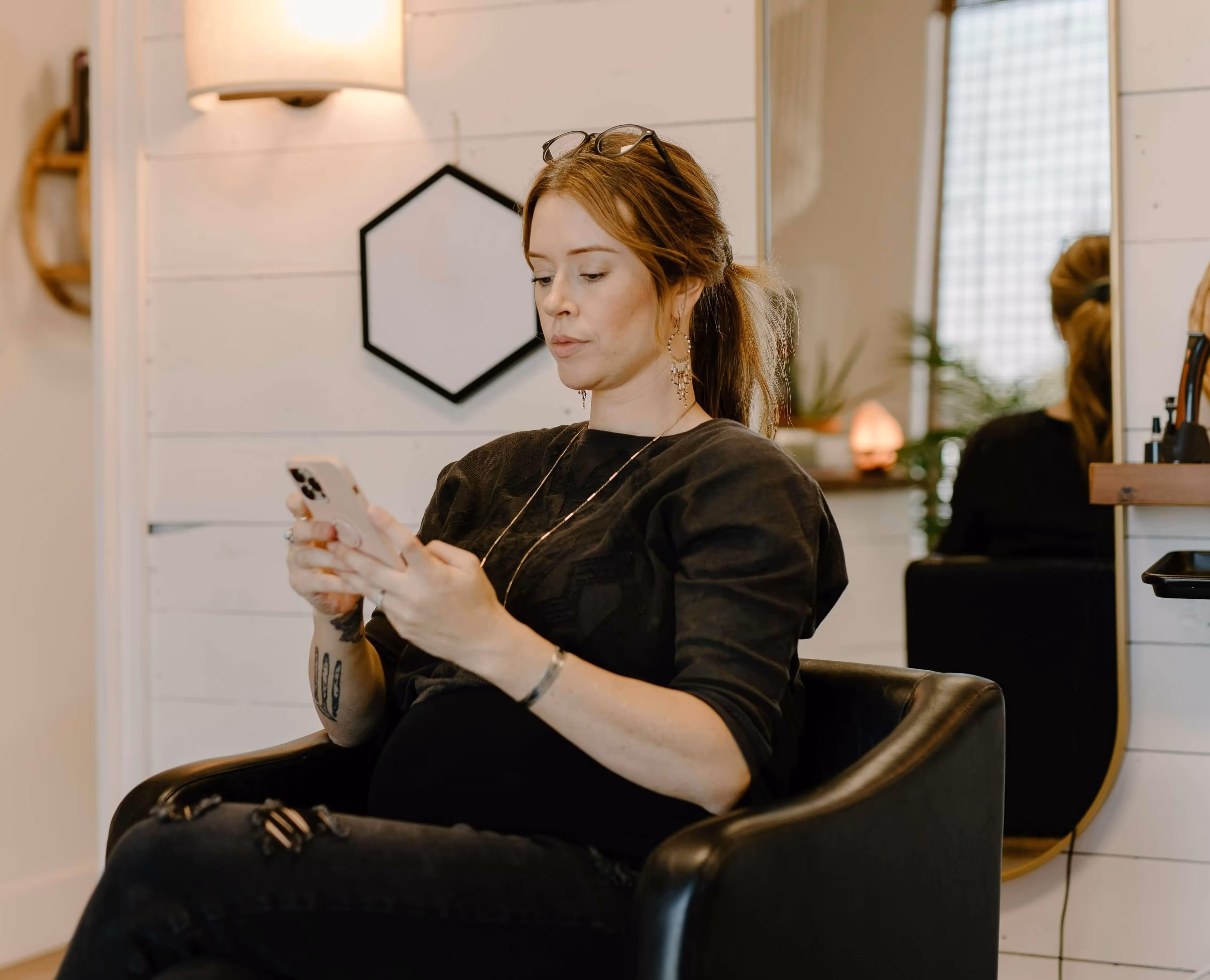 Woman with glasses on her head sitting in a black chair, looking at her smartphone in a room with a mirror and wall lamp.
