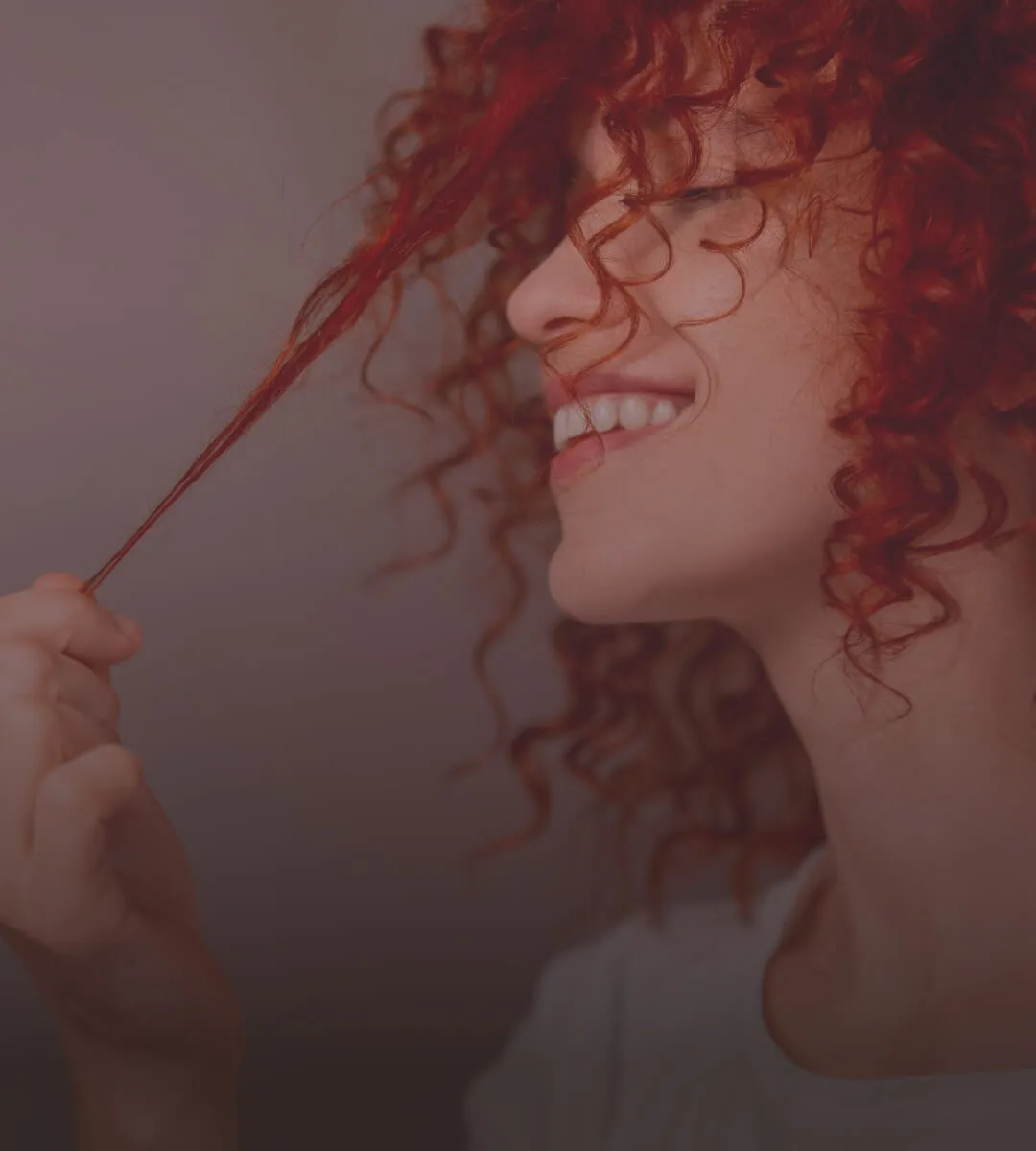 Person with curly red hair smiling and holding a strand of hair between fingers.