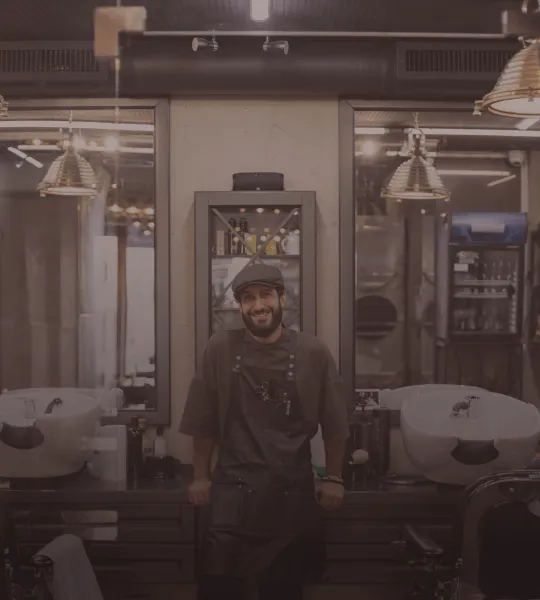 A smiling barber wearing a cap and apron standing in a vintage-style barbershop with two washbasins and hanging lights.