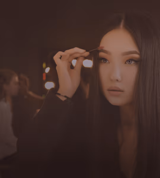 Young woman applying makeup to her eyebrows with a brush in a dimly lit room.