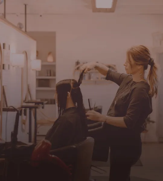 Hairdresser styling a client's long dark hair in a salon with illuminated mirrors.
