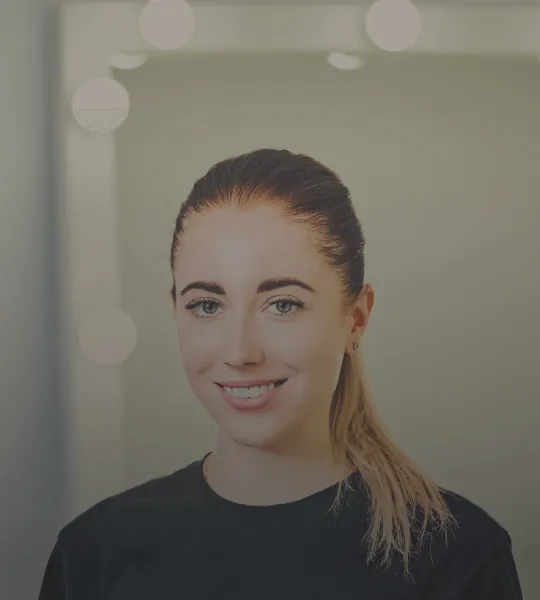 Smiling young woman with light makeup and hair tied back, wearing a black shirt in front of a blurred background with round lights.