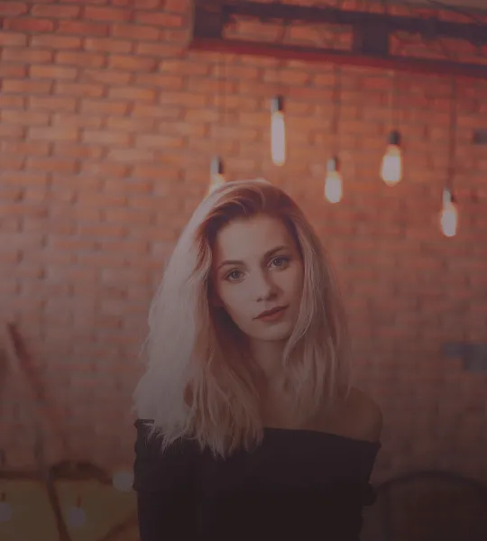 Young woman with long blonde hair wearing a black off-shoulder top standing in front of a brick wall with hanging exposed light bulbs.