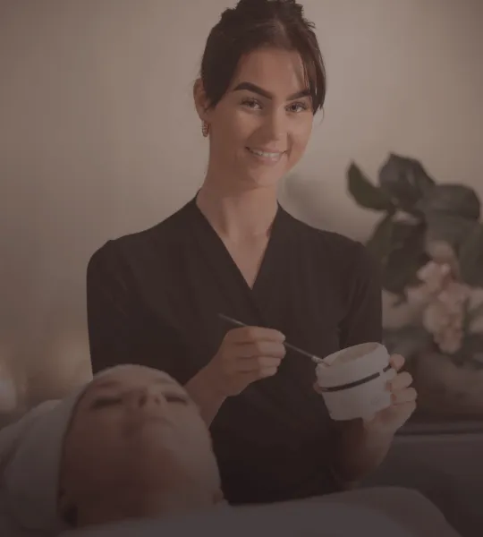 Smiling esthetician holding a jar and spatula while preparing for a facial treatment with a client lying down.