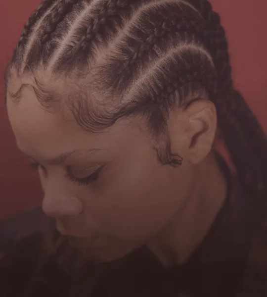 Close-up of a person with neatly braided cornrows hairstyle against a reddish background.