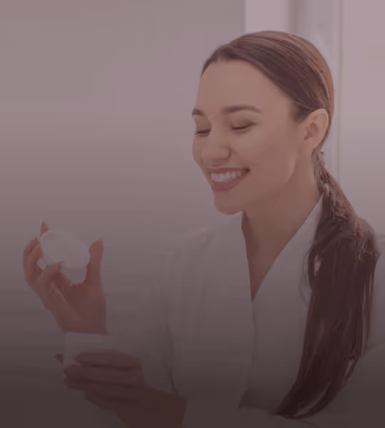 Smiling woman with long hair in a white robe holding skincare product containers.