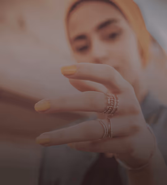 Close-up of a person's hand with yellow nail polish and multiple rings, with the person's blurred face in the background.