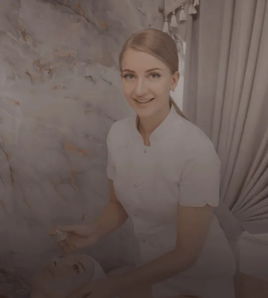 Smiling beautician in white uniform performing a facial treatment on a client lying down.