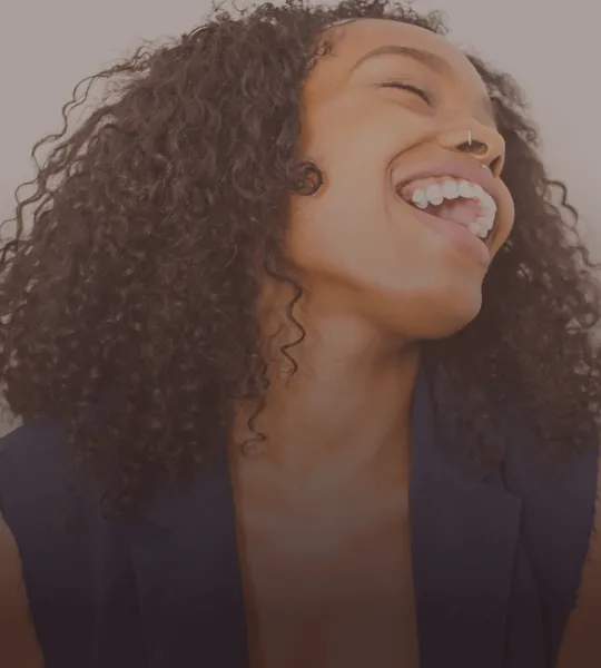 Close-up of a woman with curly hair laughing joyfully, wearing a dark blazer and a nose ring.