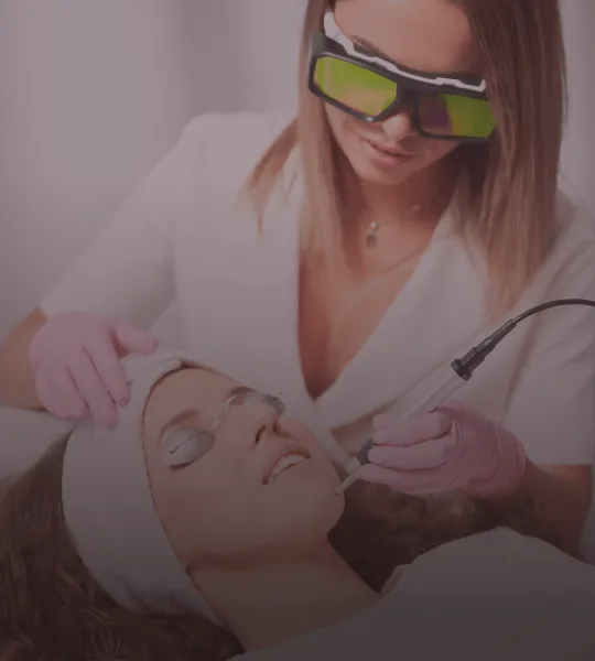 A skincare professional wearing protective glasses and gloves performing a facial laser treatment on a woman lying down with eye protection.