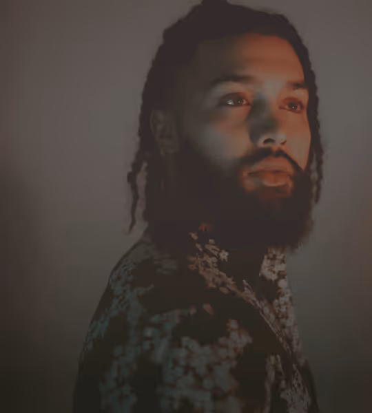 Portrait of a man with braided hair and a beard wearing a floral patterned shirt against a dark background.