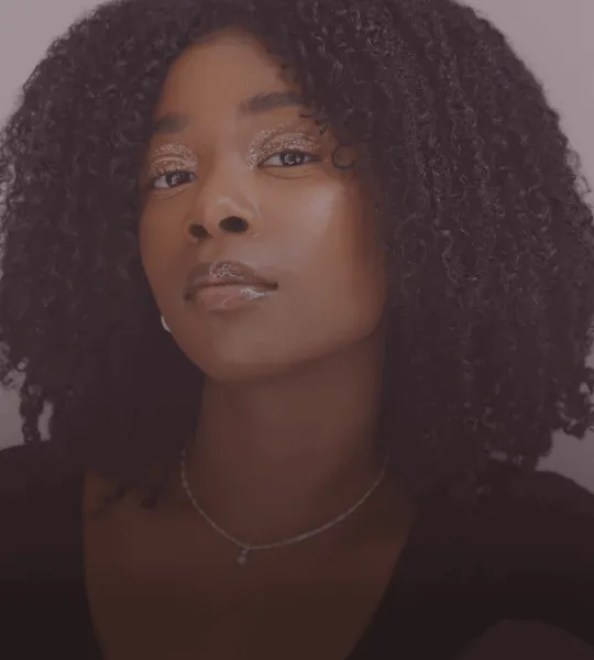 Close-up of a woman with curly hair, wearing subtle jewelry and glittery eye makeup, looking confidently at the camera.