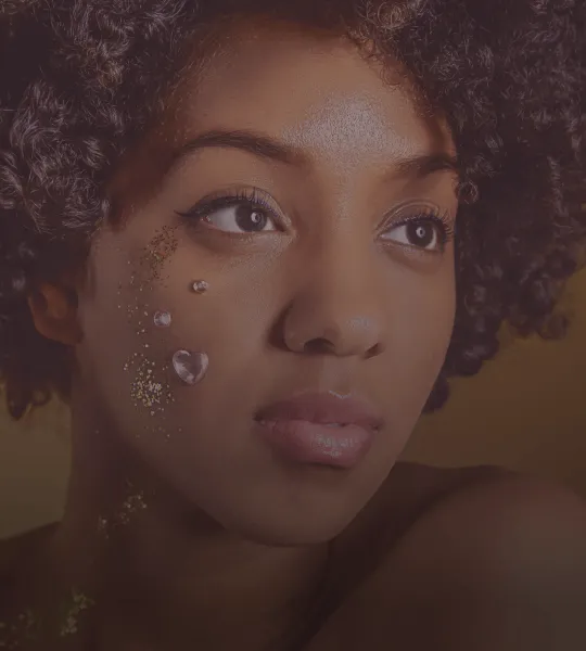Close-up of a woman with curly hair adorned with decorative gems and glitter on her cheek, looking thoughtfully to the side.
