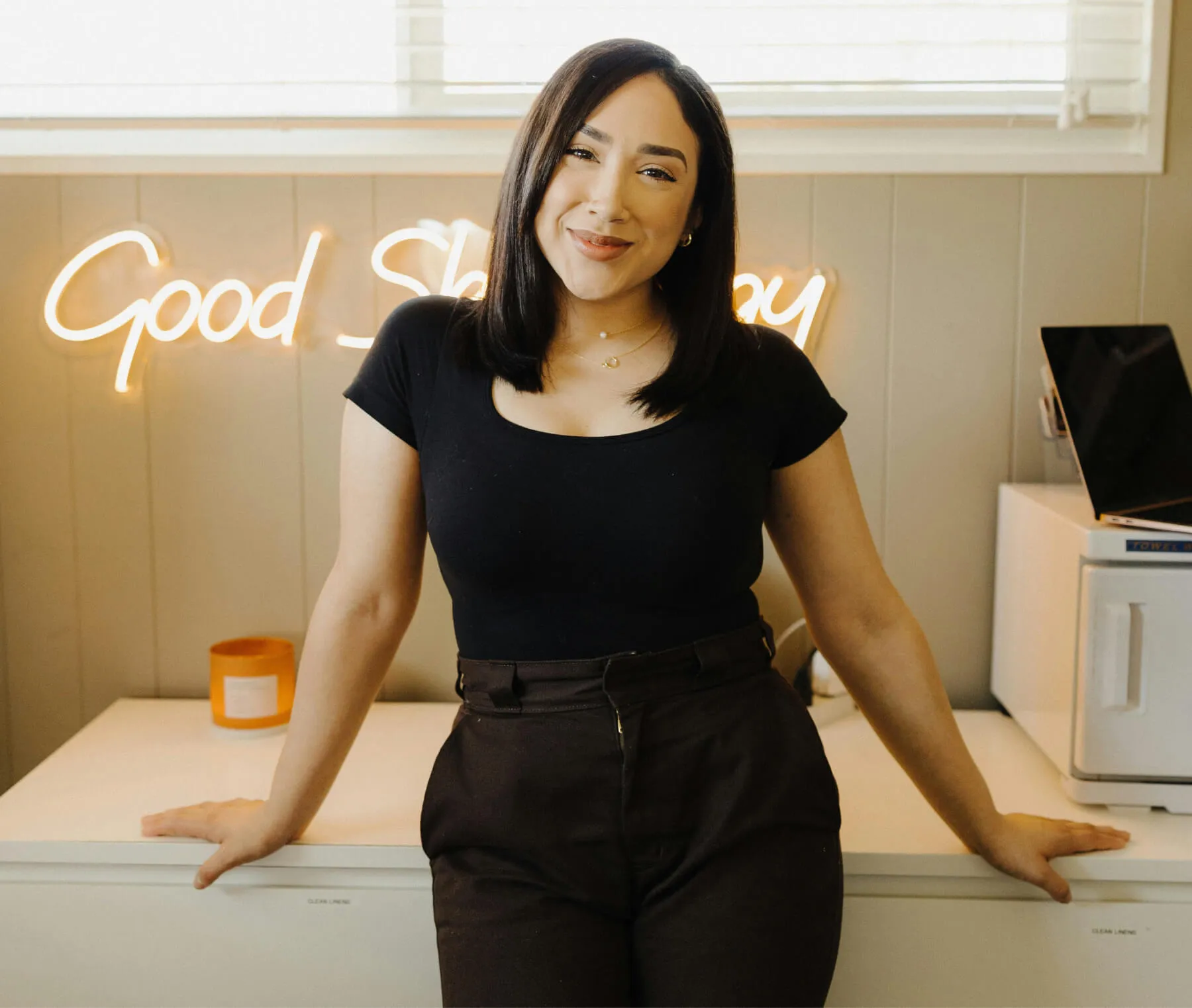 Smiling woman in black top and brown pants leaning on a white counter with a neon sign that reads 'Good Sh' in the background.