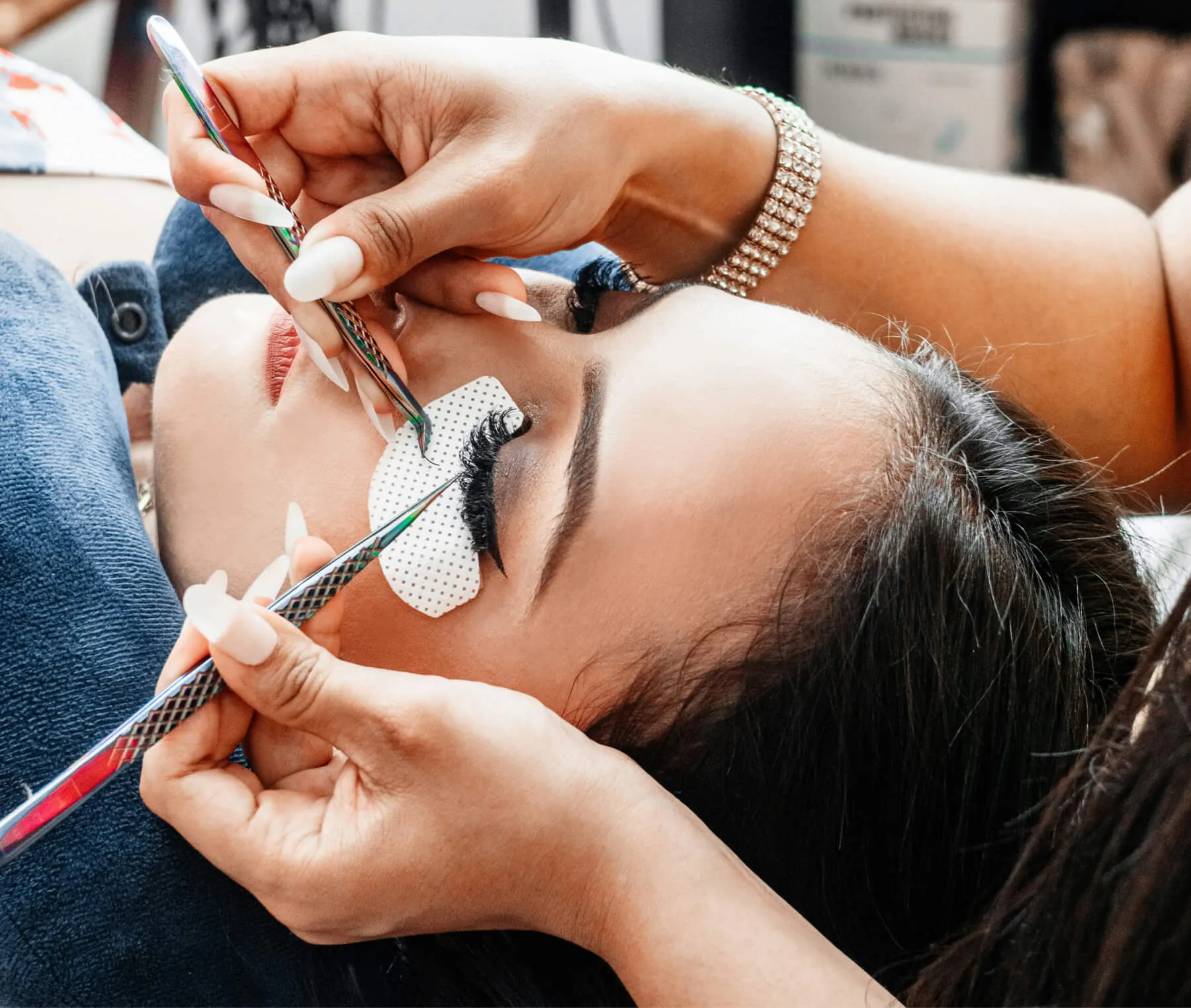 Close-up of a woman receiving eyelash extensions with a technician applying individual lashes using tweezers.
