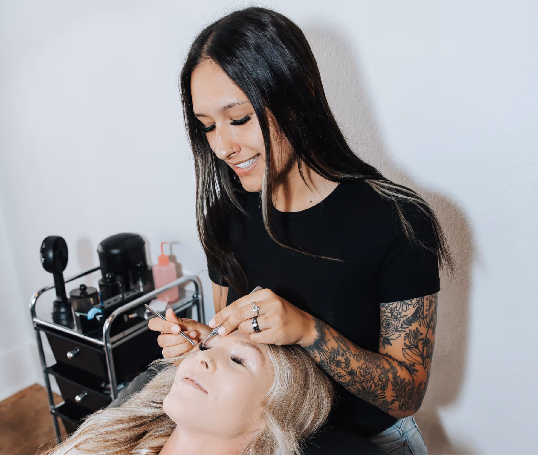 Tattooed woman applies eyelash extensions to a reclining blonde woman in a beauty salon.
