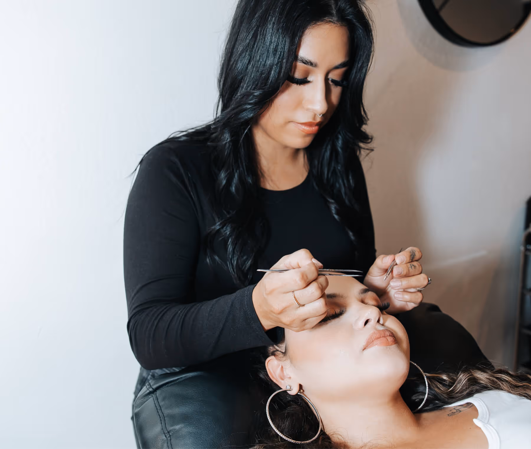 Beauty technician applying eyelash extensions to a relaxed client lying back with eyes closed.