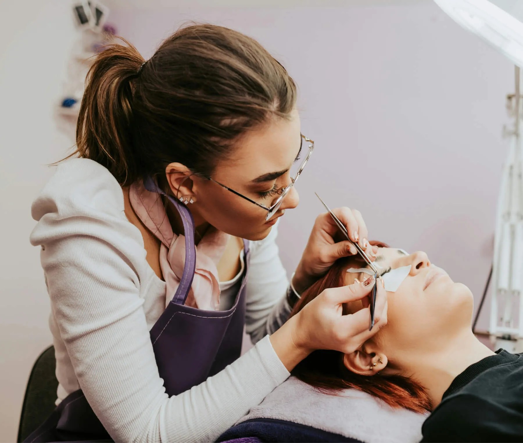 Lash technician with glasses applying eyelash extensions to a client lying down.