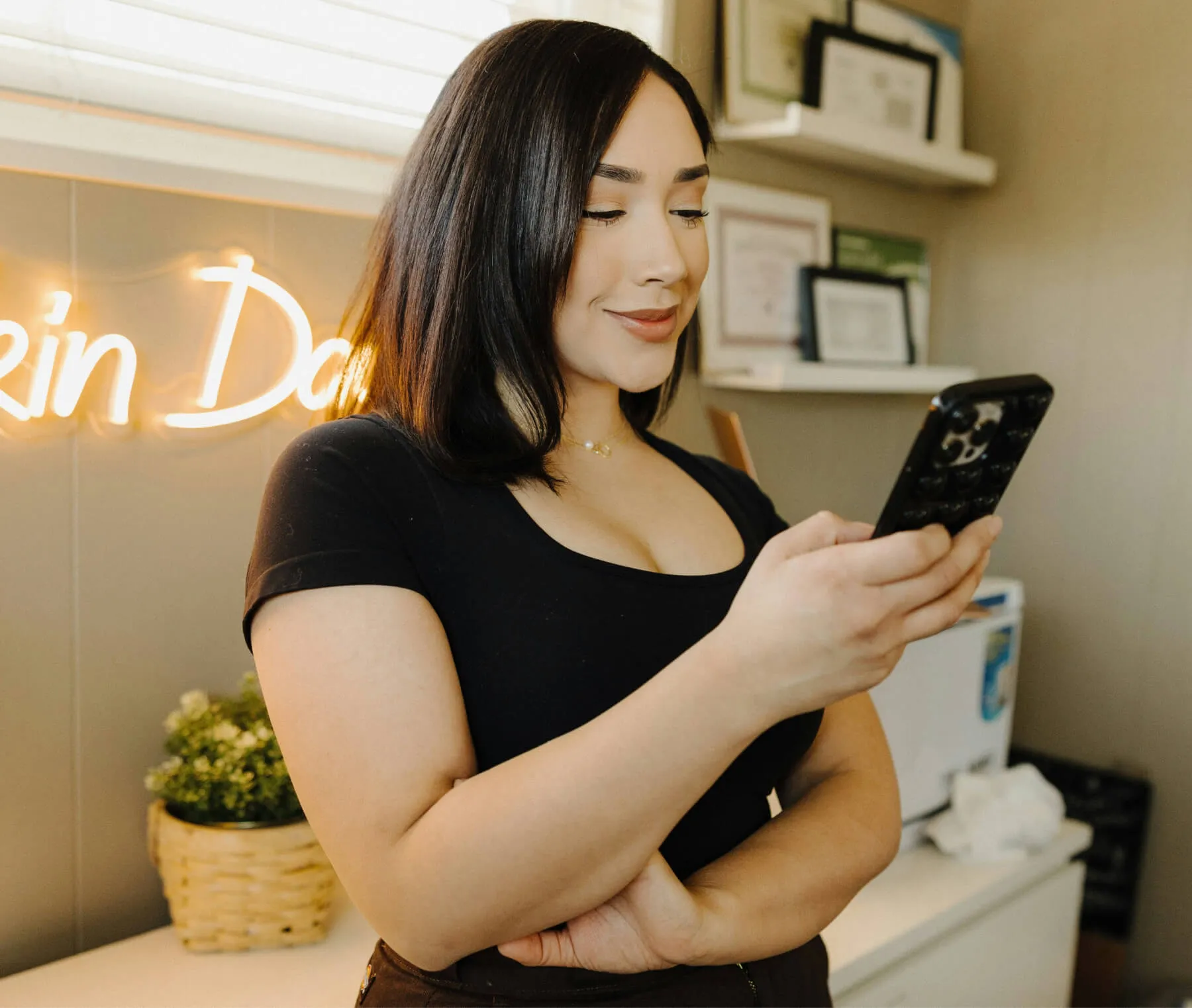 Woman smiling while looking at her smartphone in a cozy room with framed certificates and a neon sign in the background.