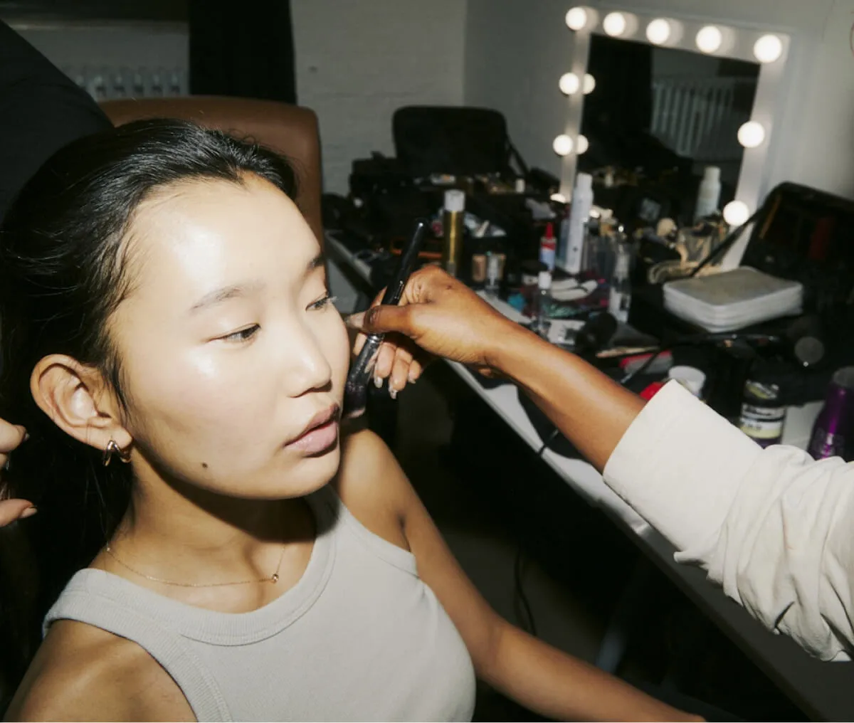 A makeup artist applying makeup with a brush on a young woman sitting near a vanity mirror surrounded by cosmetic products.