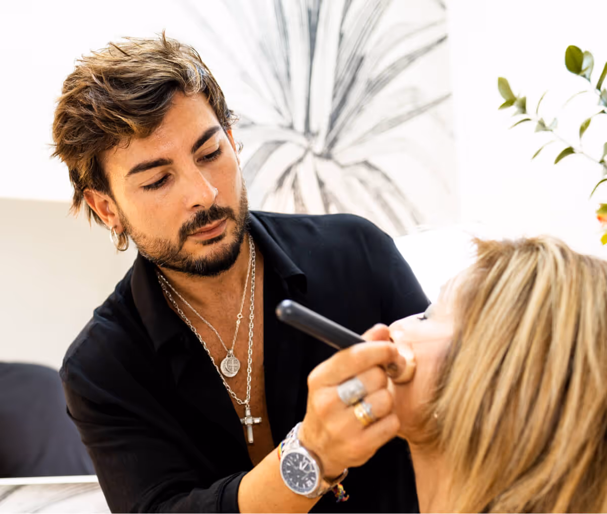 Man with beard and jewelry applying makeup on a blonde woman indoors.