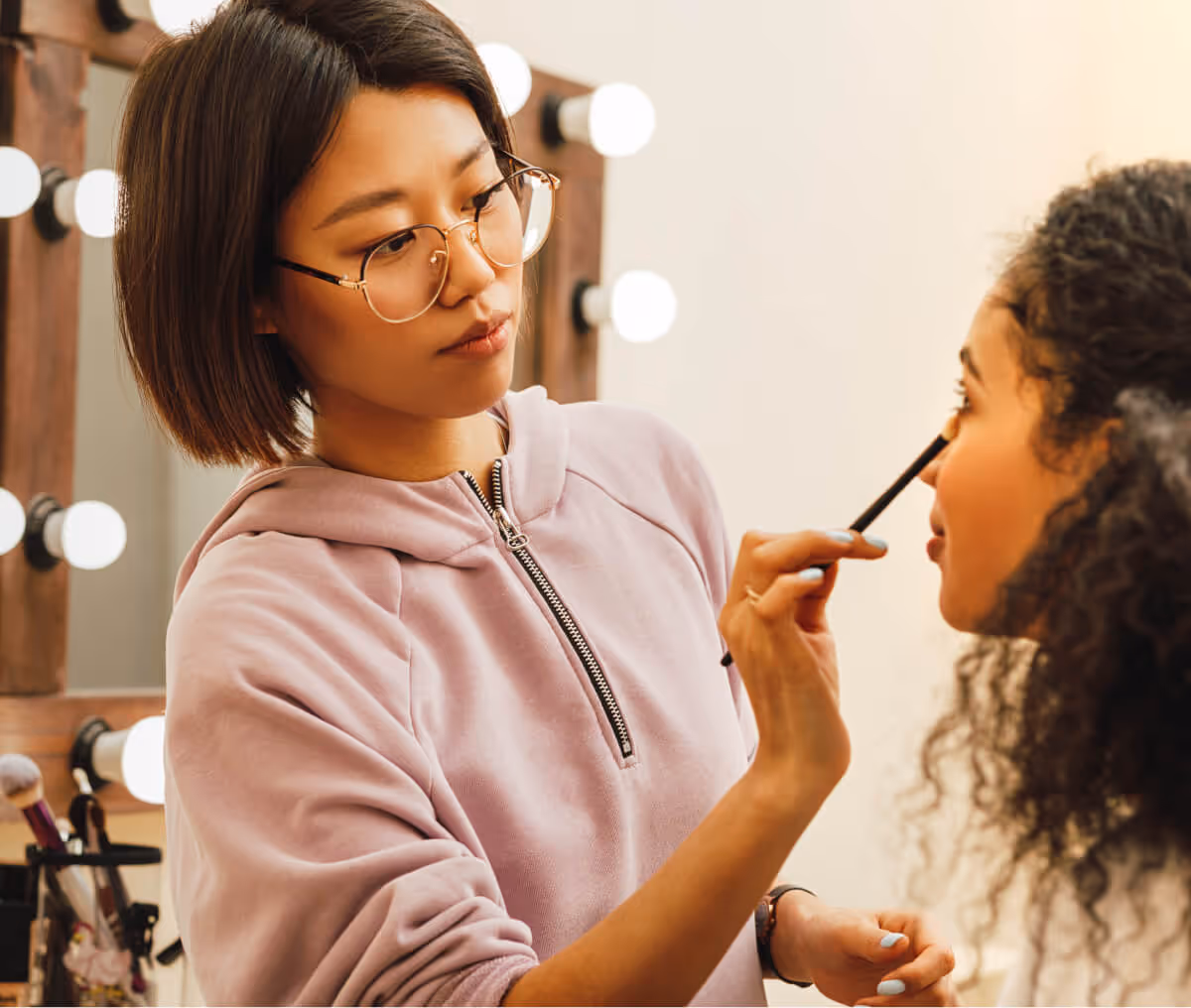 Makeup artist wearing glasses and a pink hoodie applying eyeshadow to a woman with curly hair in a lighted vanity mirror setting.