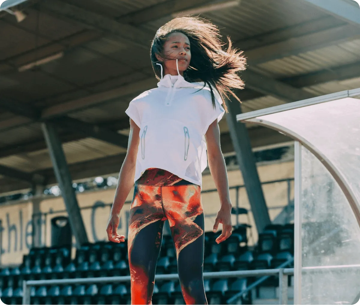 Young woman with flowing hair wearing a white sleeveless hoodie and red patterned leggings standing at an outdoor stadium.