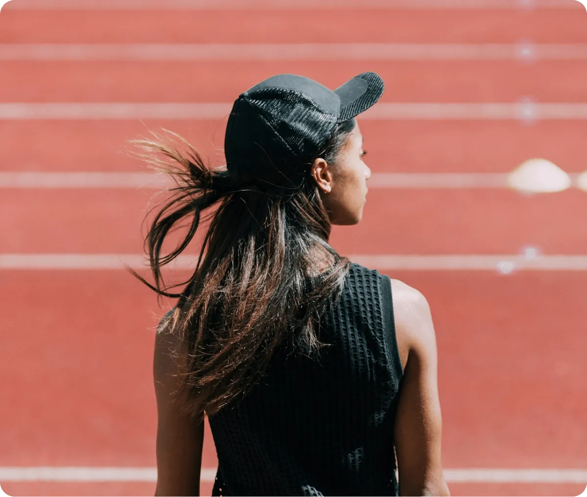 Woman in black sleeveless top and cap standing with her back to the camera on a red running track.