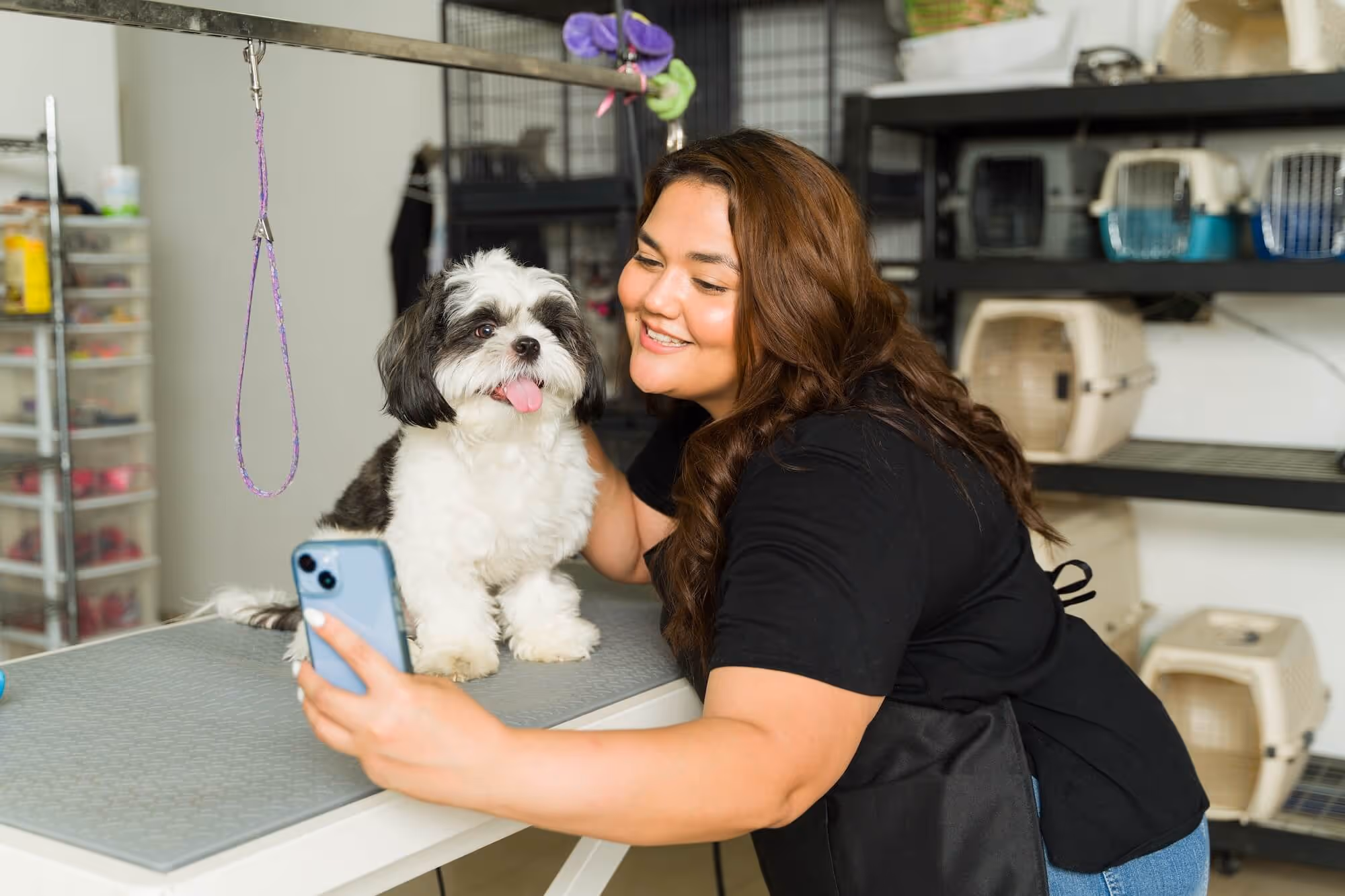 Woman taking a selfie with a small black and white dog on a grooming table in a pet grooming salon.