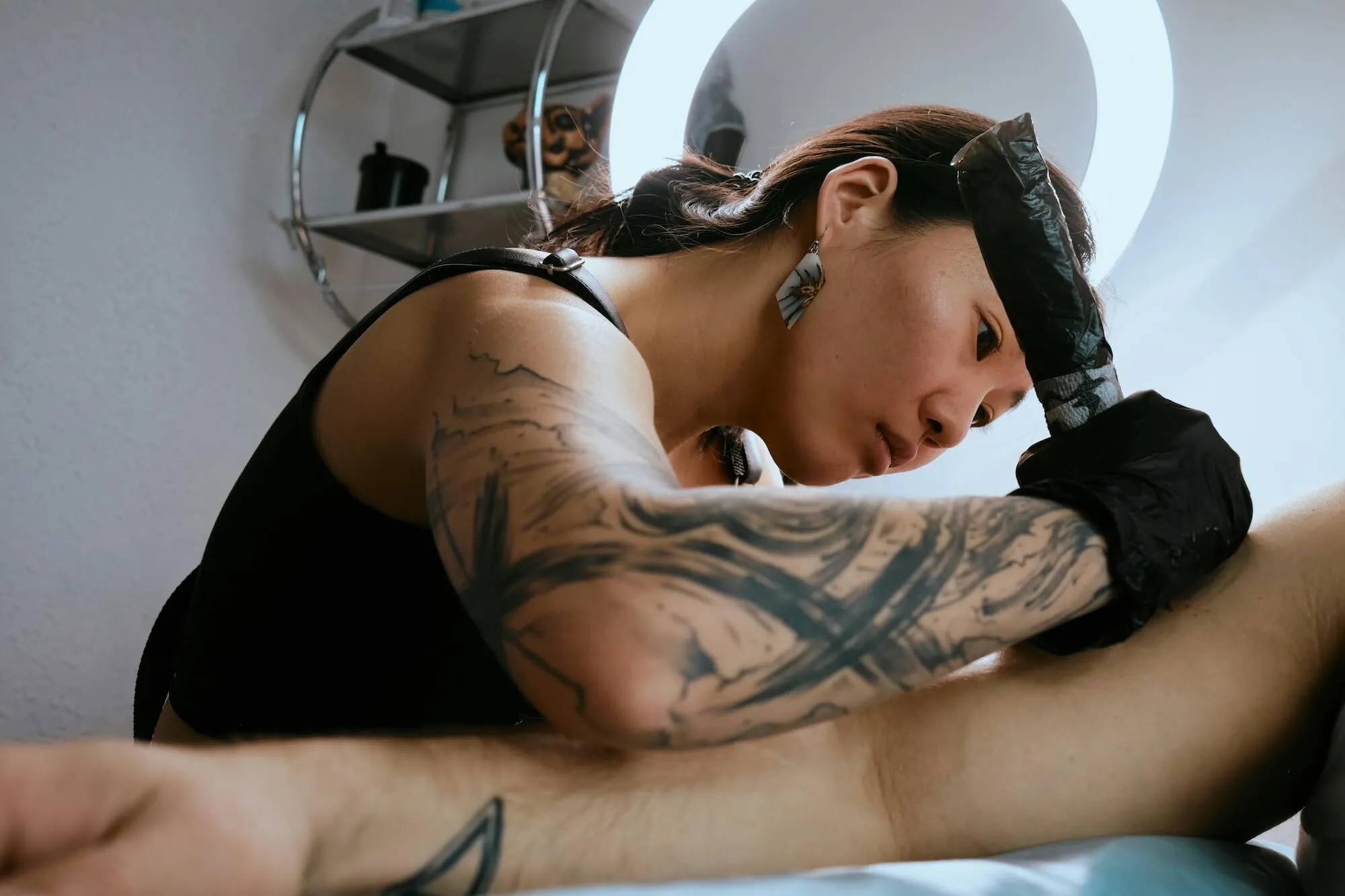Tattoo artist with tattooed arm and gloves focused on tattooing a client's arm under a ring light.