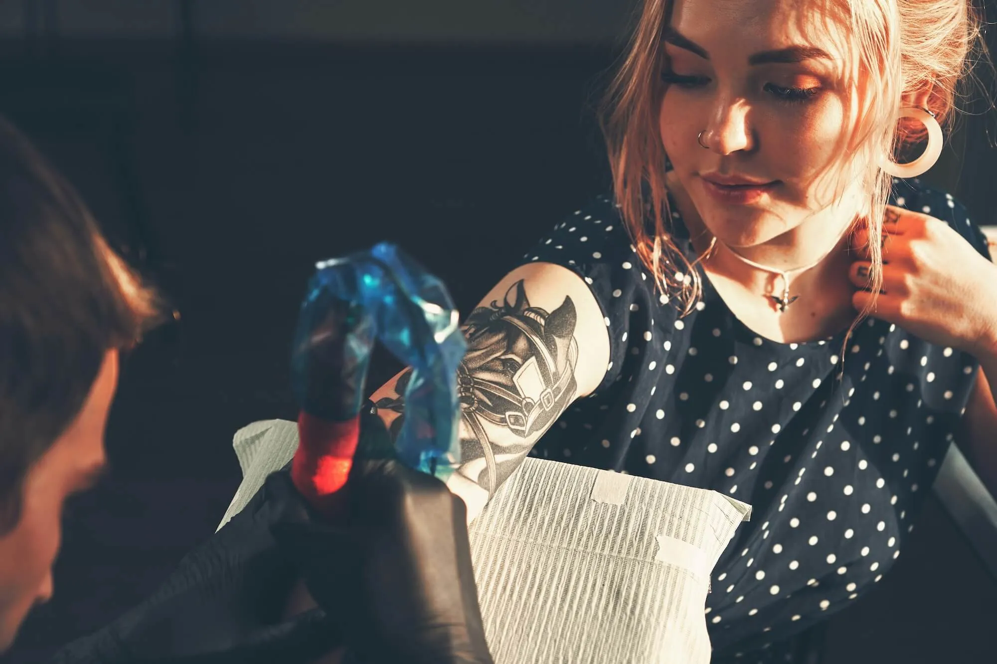 Tattoo artist wearing black gloves working on a detailed black and gray tattoo of a horse head on a woman's upper arm.